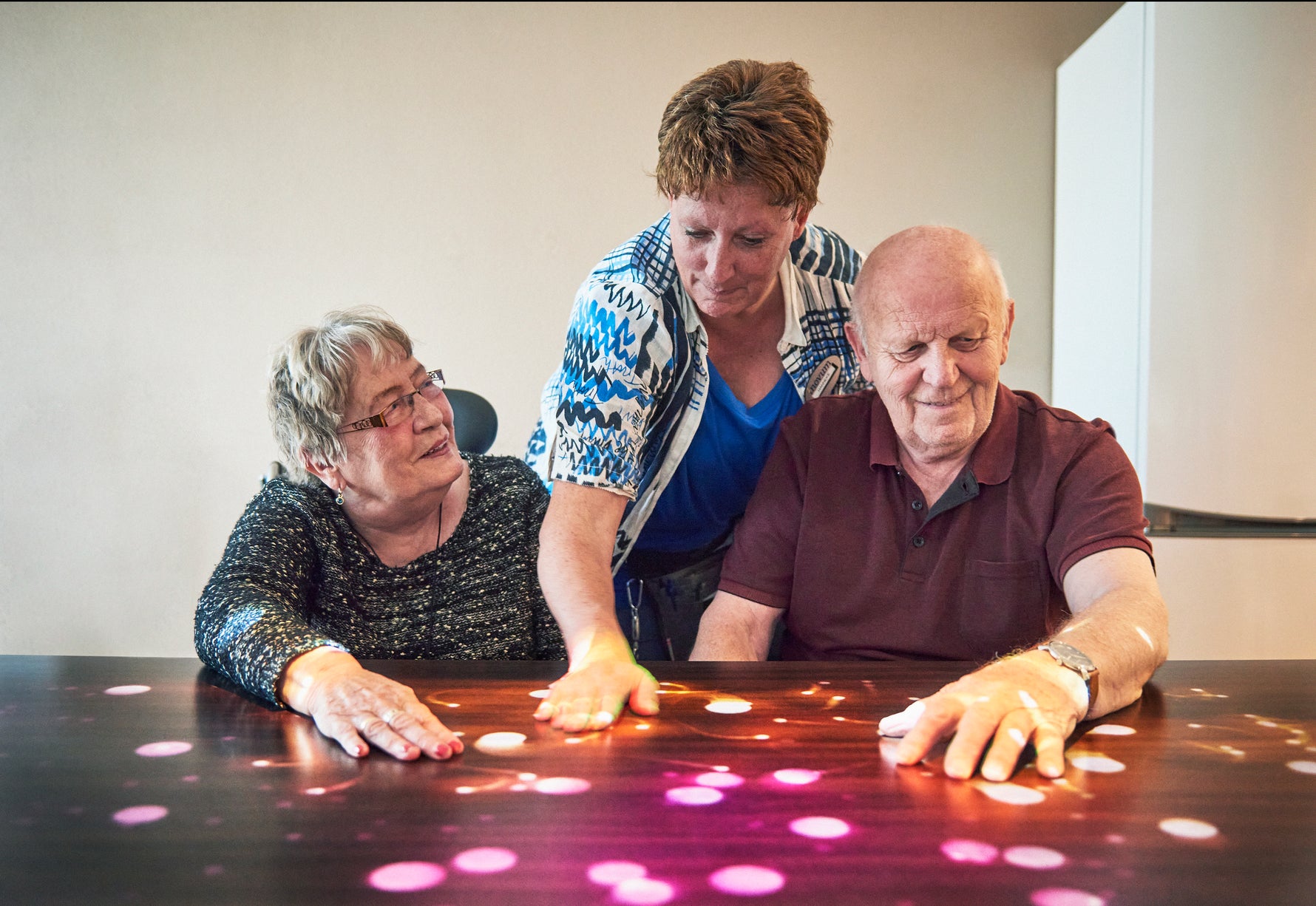 Three people interacting with the Tovertafel, which projects a glowing pink and white pattern on a table