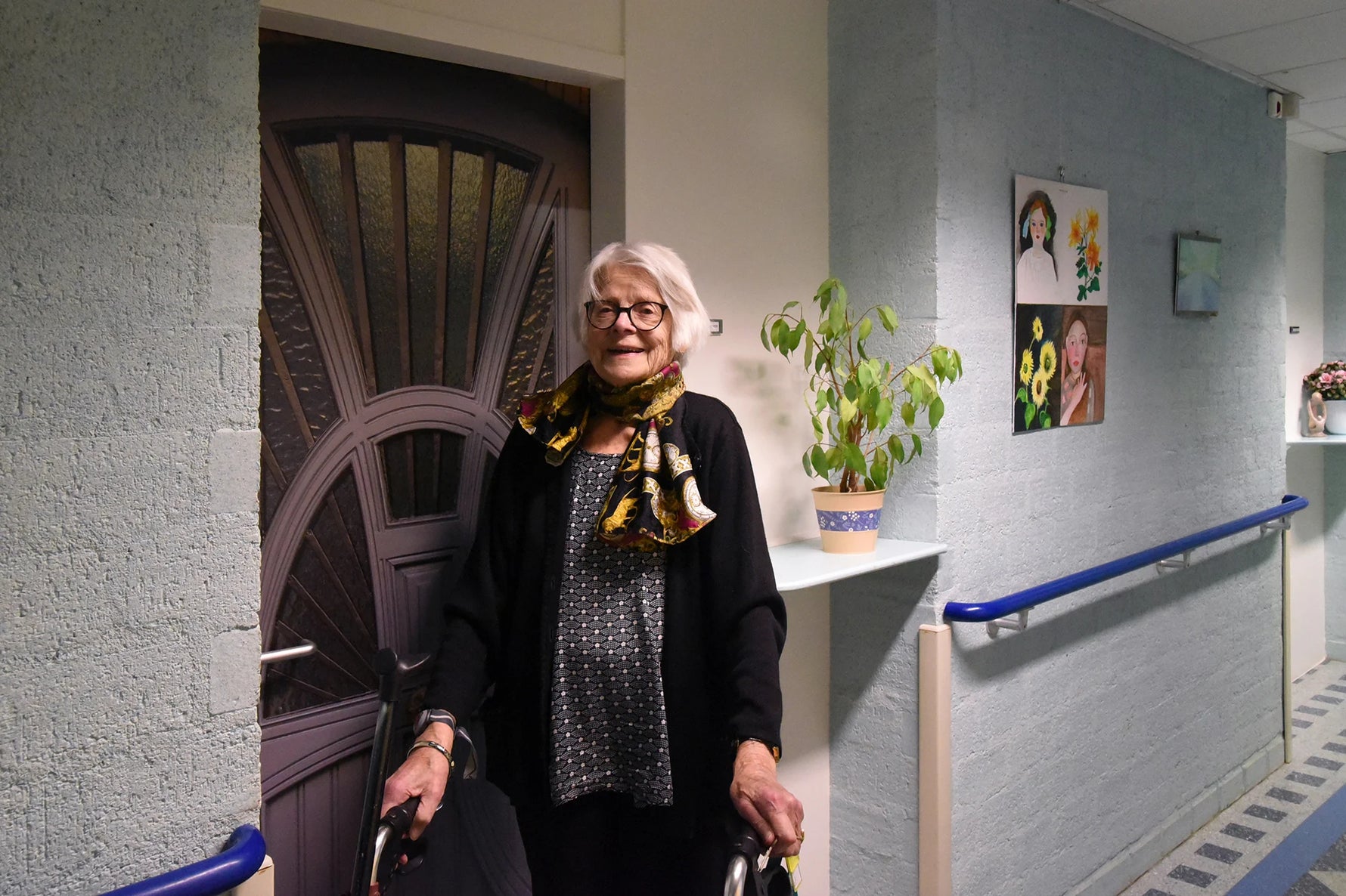 Woman standing in a hallway with a walker, wearing glasses and a patterned scarf. She's standing in front of her True Doors door.