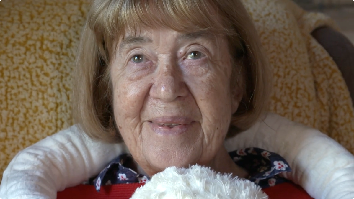 An elderly woman with short blonde hair, wearing a white sweater over a patterned shirt, sitting on a couch.