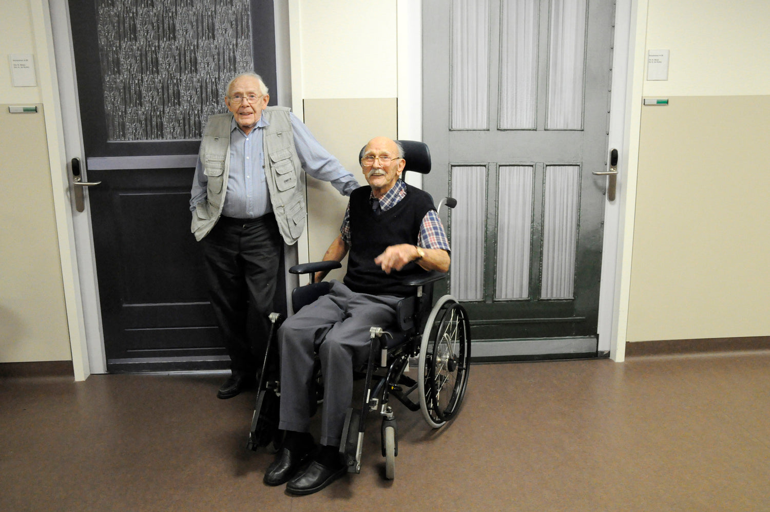 Two elderly men, on standing and the other in a wheelchair, smiling in front of their True Doors decal doors.