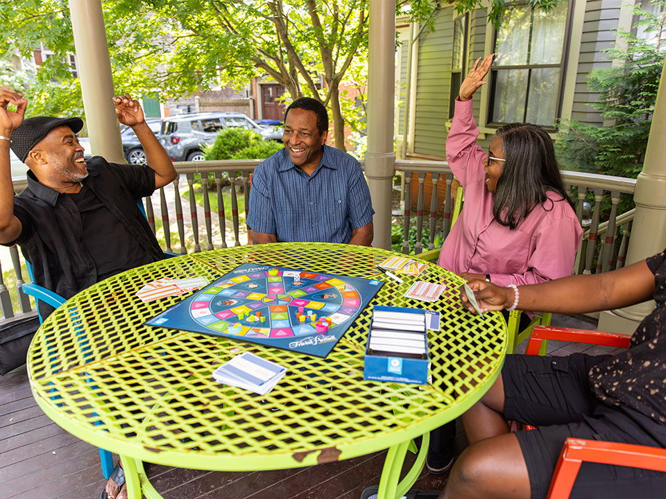 A group older adults smiling and playing Trivial Pursuit generations outside on their patio table.