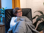 Lady sitting in a sofa chair curled up in the Fiora weighted blanket.