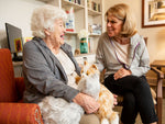 An elderly woman sitting with her caregiver in a living room holding both the silver and the orange cat.