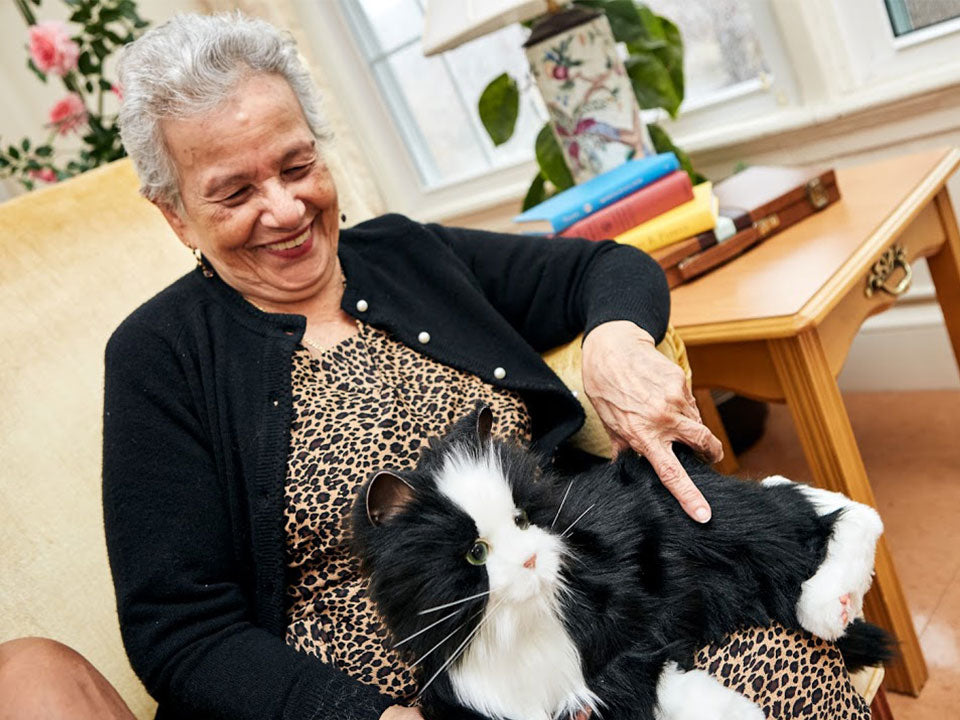 Older woman, smiling as she pets the tuxedo Joy for All interactive companion cat.